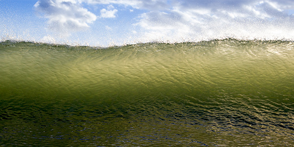 A green wave crashing, highlighted against a blue sky with clouds.