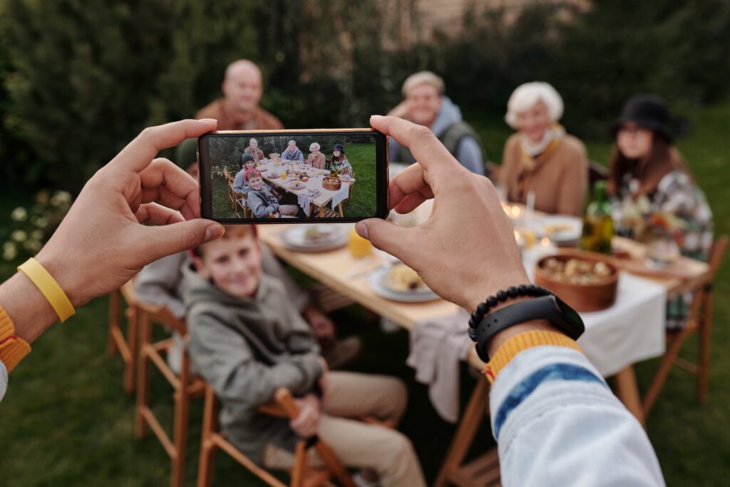 A multi generational family is photographed with a smart phone outside enjoying a picnic